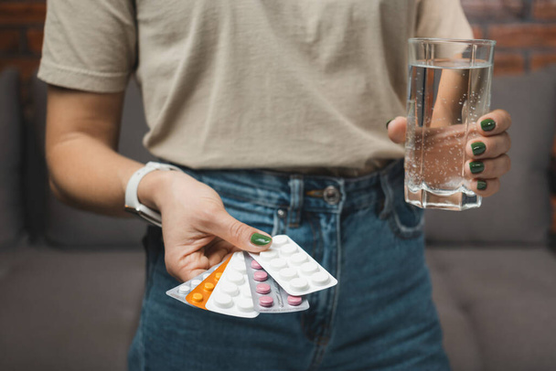 Young woman holds a medicine, various blister packs with pills for treatment and glass of water, close-up view. - Photo, Image