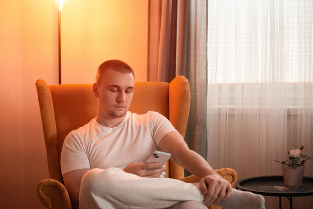 Young adult man freelancer using his mobile phone checking social media networks feed or message chat sitting on cozy chair at home. Online digital communication, rest after hard work day - Photo, Image