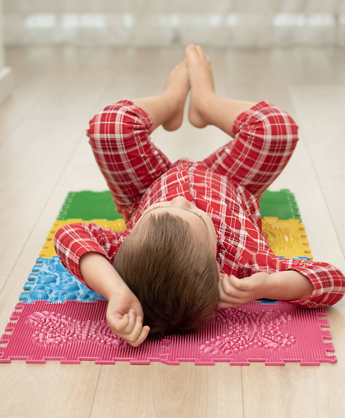 Concepto de deporte y salud. Un niño de 4 años en pijama a cuadros rojo se está divirtiendo en una alfombra ortopédica de masaje multicolor con púas en el interior de una casa. Primer plano. Enfoque suave. - Foto, imagen