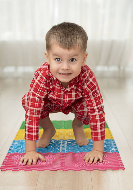 Sport- und Gesundheitskonzept. Ein kleiner Junge von 4 Jahren im rot karierten Pyjama amüsiert sich auf einer mehrfarbigen Massagematte mit Spikes im häuslichen Innenraum. Nahaufnahme. Weicher Fokus. - Foto, Bild