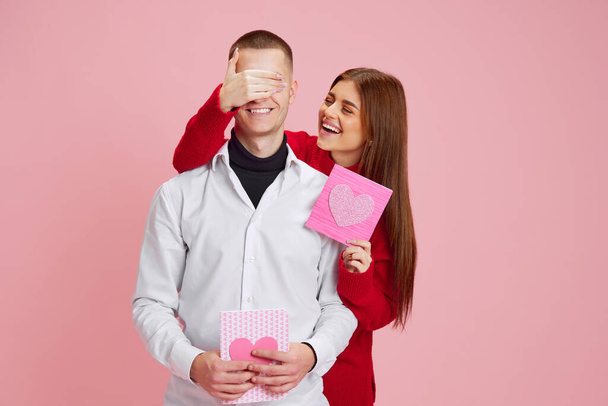 Young girl covering boyfriends eyes and presenting him little gift, postcard with heart against pink studio background. Concept of love, relationship, Valentines Day, emotions, lifestyle - Photo, Image