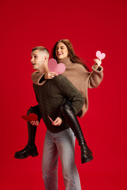Playful, young, happy couple, man and woman, boyfriend and girlfriend having fun against red studio background. Concept of love, relationship, Valentines Day, emotions, lifestyle - Fotoğraf, Görsel