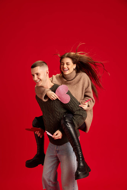 Playful, young, happy couple, man and woman, boyfriend and girlfriend having fun against red studio background. Concept of love, relationship, Valentines Day, emotions, lifestyle - Фото, зображення