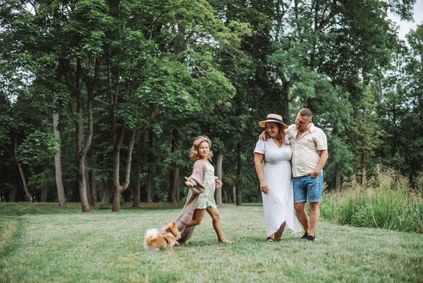 Małżeństwa ściskają się na zielonym trawniku i patrzą, jak ich córka bawi się obok psa. Rodzina chodzi na świeżym powietrzu. Harmonijna kochająca szczęśliwa rodzina. Rodzinne wakacje. Szczęśliwi razem. - Zdjęcie, obraz
