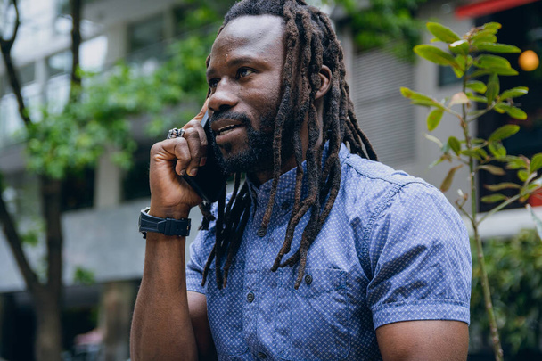 Close-up do homem africano com dreadlocks e barba, sentado ao ar livre, discutindo falar sobre telefonema, tecnologia e estilo de vida conceito, espaço de cópia. - Foto, Imagem