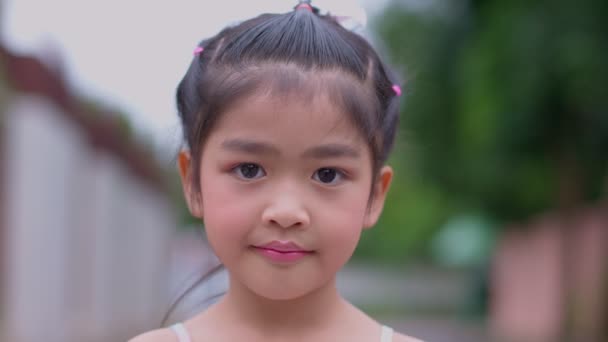 Portret van een aantrekkelijk Aziatisch meisje dat glimlacht en naar de camera kijkt en zwaait met haar hand, gelukkig lachend kind dat naar de camera kijkt - van dichtbij, buiten. Vreugdevolle natuur jeugdrecreatie concept. - Video