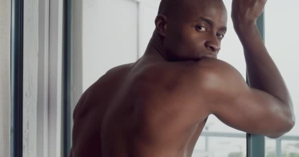 Knap, gezicht en rug van zwarte man door raam met spieren voor een zelfverzekerde houding in het appartement. Serieus, shirtloos en portret van een jonge Afrikaan in de slaapkamer van een modern huis of huis - Video