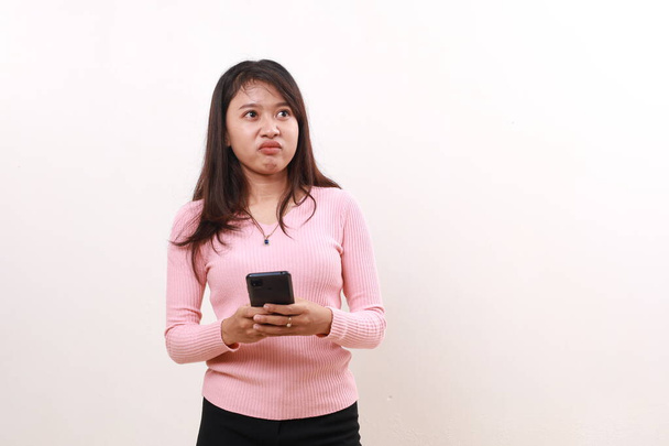 Unzufriedene junge Asiatin, die oben steht und ein Handy in der Hand hält. Isoliert auf weißem Hintergrund - Foto, Bild