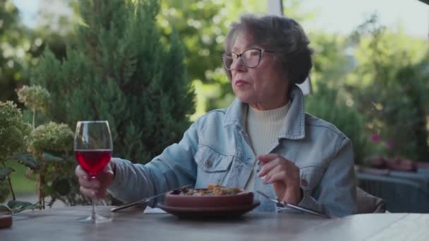 An elderly woman relaxes outdoors on a sunny autumn or spring day and looks ahead. An elegant lady sits in the city park at a table in a street cafe, enjoys the taste of delicious dish and drinks red - Footage, Video