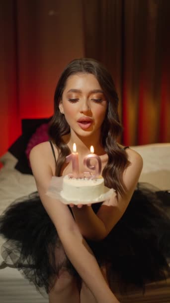 Una bella giovane bruna con una torta in mano spegne una candela in una stanza buia. Bruna sexy che spegne una candela in una stanza buia di una stanza d'albergo - Filmati, video