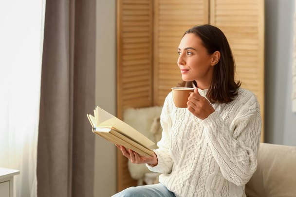 Νεαρή γυναίκα με φλιτζάνι καφέ διαβάζοντας βιβλίο στο σπίτι - Φωτογραφία, εικόνα