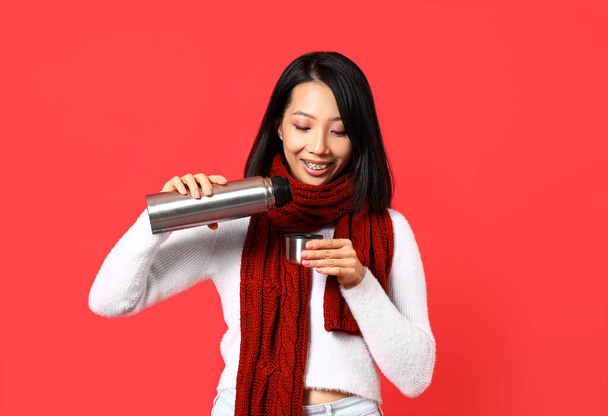 Молодая азиатка в зимней одежде с термосом на красном фоне - Фото, изображение