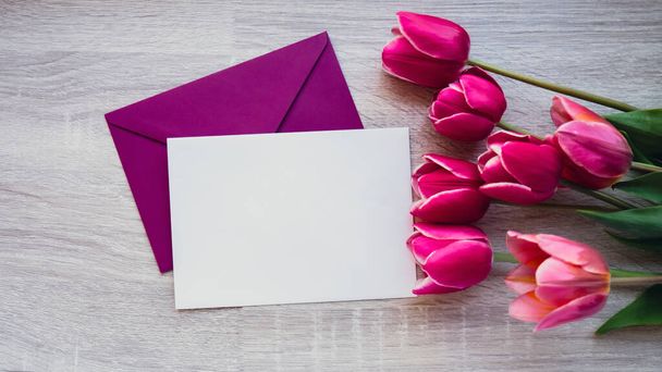 Пустой белый конверт копировать пространство для текста или дизайна с красивым розовым букетом тюльпанов на деревянном фоне. Шаблон для праздничной весенней открытки. Вид сверху Плоская пустая бумага - Фото, изображение