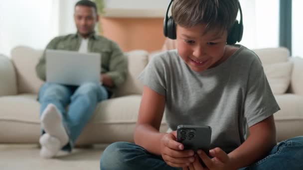 Маленький мальчик сын играет в видеоигры на мобильном телефоне в наушниках на полу счастливой многорасовой семьи с использованием различных устройств дома мама папа работает в Интернете на ноутбуках фон ребенка играть смартфон - Кадры, видео