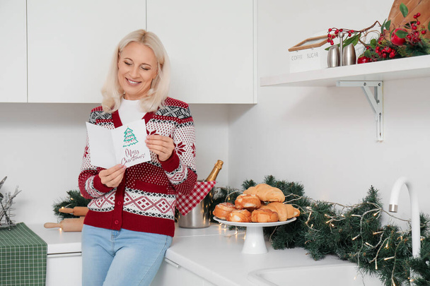 Ώριμη γυναίκα με ευχετήρια κάρτα για Χριστουγεννιάτικη γιορτή στην κουζίνα - Φωτογραφία, εικόνα