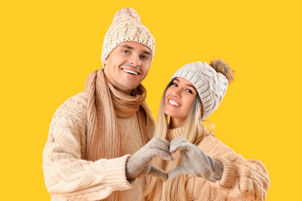 Glückliches junges Paar in Winterkleidung macht Herzensgeste auf gelbem Hintergrund - Foto, Bild