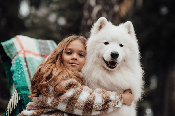 Weibliche Kinder feiern Weihnachten und Neujahr im Freien. Kleines Mädchen verbringt fröhliche Zeit mit weißem Samojeden-Hund in der Nähe des Weihnachtsbusses und genießt die Kindheit - Foto, Bild