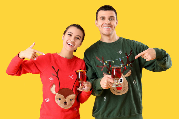 Young couple in Christmas sweaters pointing at mulled wine on yellow background - Photo, Image