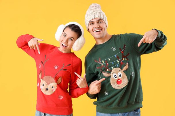 Young couple pointing at Christmas sweaters on yellow background - Photo, Image