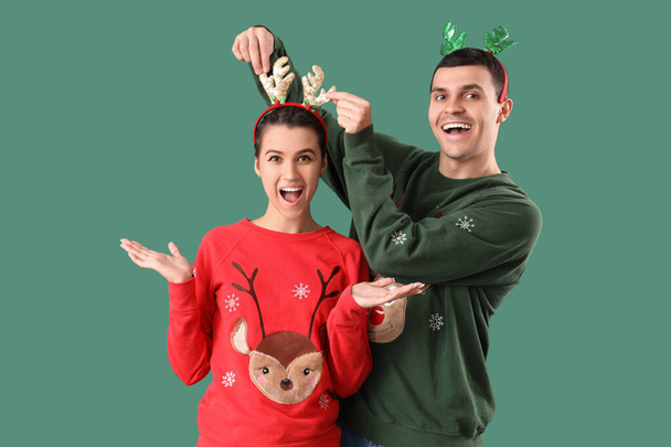 Young couple in Christmas sweaters and reindeer horns on green background - Photo, Image