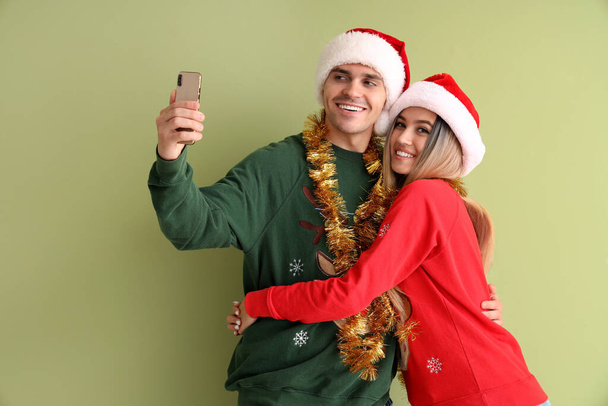Young couple in Santa hats and Christmas sweaters taking selfie on green background - Photo, Image