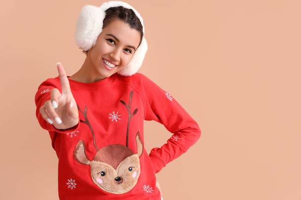 Junge Frau in Weihnachtspullover und Ohrenschützer zeigt auf etwas auf beigem Hintergrund - Foto, Bild