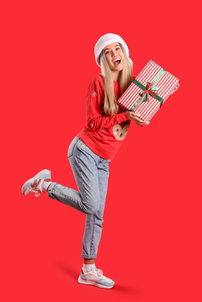 Young woman in Santa hat and Christmas sweater with gift on red background - Photo, Image