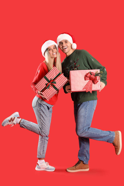 Young couple in Santa hats with Christmas gifts on red background - Photo, Image