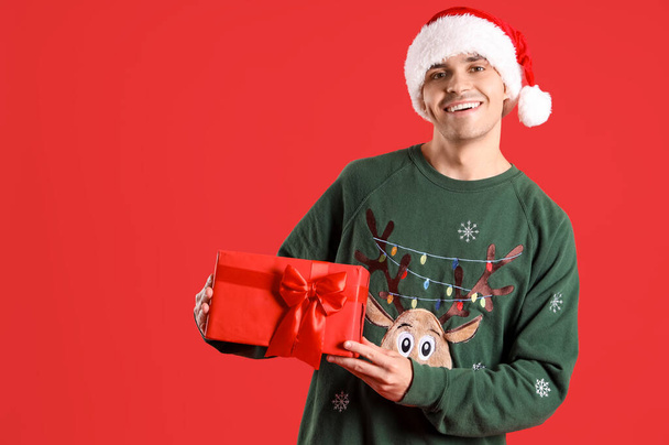 Young man in Santa hat and Christmas sweater with gift on red background - Photo, Image