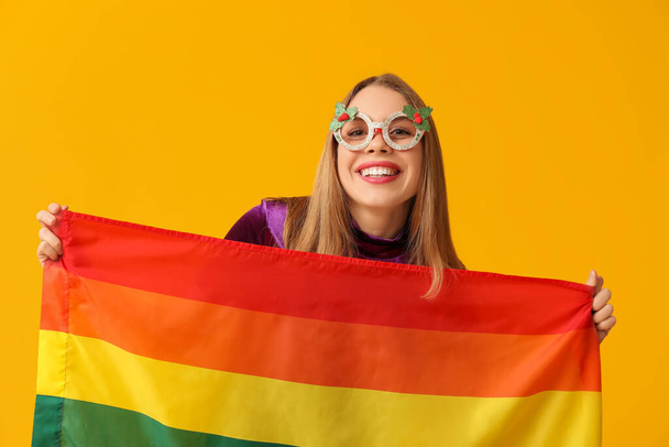 Young woman in Christmas eyeglasses with LGBT flag on yellow background - Photo, Image