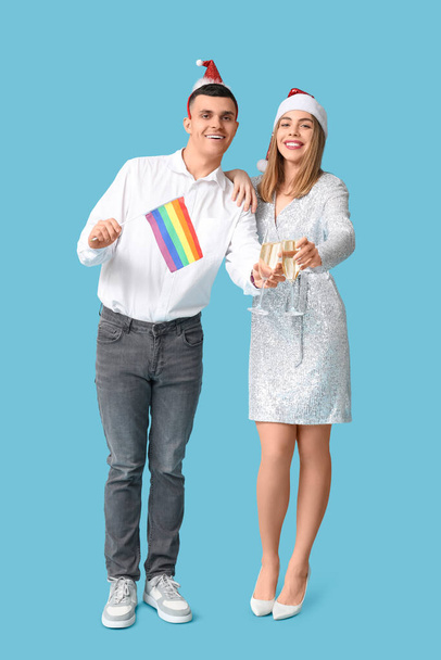 Молода пара в Санта-капелюхах з прапором ЛГБТ та шампанським на синьому фоні - Фото, зображення