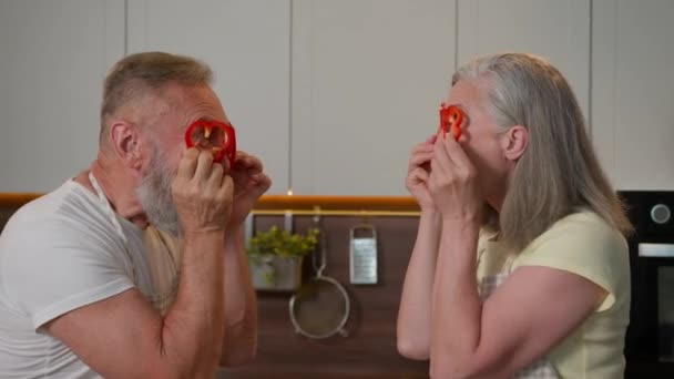 Αστεία ευτυχισμένη συνταξιούχος γιαγιά παππούς Καυκάσιος γέρος γυναίκα διασκεδάζοντας μαζί κάνουν κόκκινο γυαλιά πάπρικα κύκλους πιπεριές μεσήλικες σύζυγος ηλικιωμένοι παππούδες μαγείρεμα φρέσκα λαχανικά στην κουζίνα - Πλάνα, βίντεο