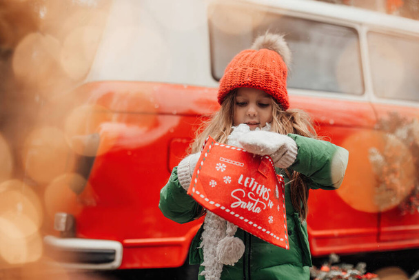 Weibliche Kinder feiern Weihnachten und Neujahr im Freien. Kleines Mädchen verbringt freudig Zeit im Freien mit Brief an den Weihnachtsmann in rotem Umschlag und wartet auf ein Wunder - Foto, Bild