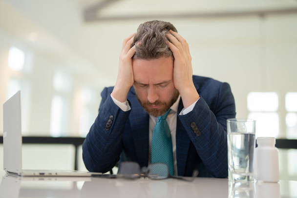 Un homme d'affaires stressé a mal à la tête au bureau. Homme d'affaires fatigué fait des heures supplémentaires et a mal à la tête. Homme surmené avec ordinateur portable sur le lieu de travail, souffre de maux de tête. Migraine du surmenage - Photo, image