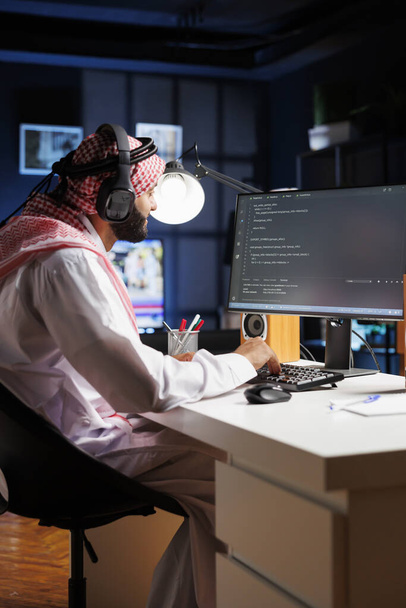 Разработчик данных с Ближнего Востока пишет код на компьютере, демонстрируя ядро программирования и разработки программного обеспечения. Мусульманин, использующий беспроводные наушники и погруженный в выполнение алгоритмов. - Фото, изображение
