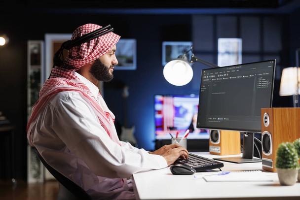 Η εικόνα δείχνει έναν Άραβα προγραμματιστή λογισμικού που εργάζεται στην οθόνη του υπολογιστή, καθισμένος σε έναν αλγόριθμο επεξεργασίας θέσης εργασίας. Ο μουσουλμάνος κωδικοποιητής χρησιμοποιεί έναν επιτραπέζιο υπολογιστή για να εργαστεί στη διεπαφή χρήστη. - Φωτογραφία, εικόνα