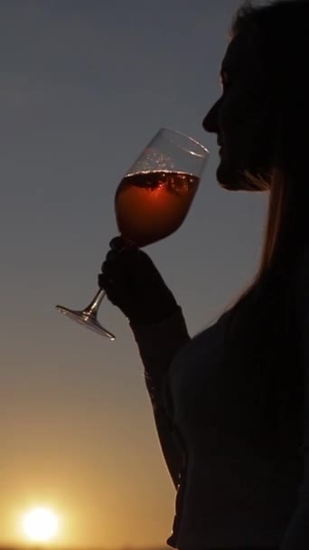 Η σιλουέτα μιας γυναίκας που πίνει κρασί από ένα ποτήρι το ηλιοβασίλεμα. Κάθετη βίντεο. - Πλάνα, βίντεο