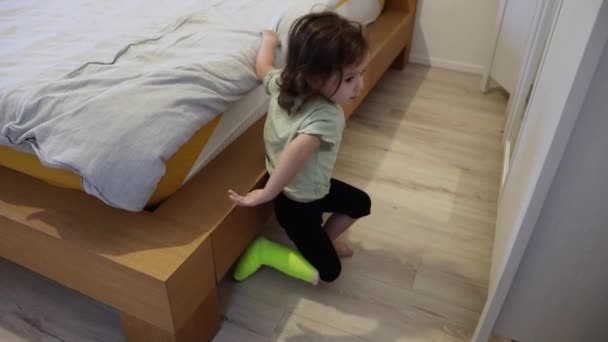 Una pequeña chica caucásica hermosa con un elenco verde claro en su pierna izquierda se levanta de la cama grande en el dormitorio en el suelo, se arrodilla y va a jugar con los juguetes, vista lateral de cerca. - Metraje, vídeo