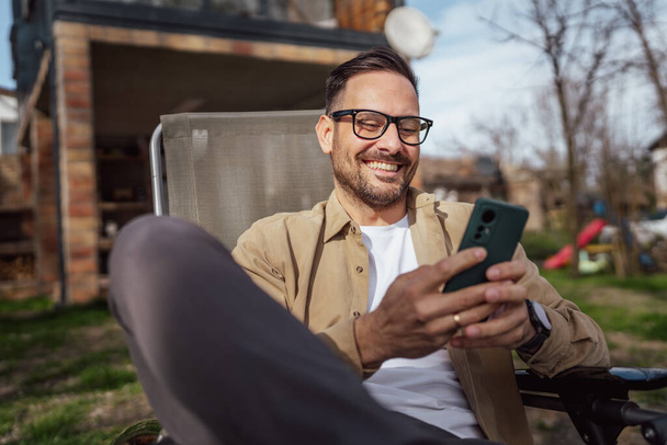 Ein Mann kaukasischen Mann mit Bart und Brille sitzen im Freien in sonnigen Tag tragen Hemd verwenden Handy für SMS-Nachrichten surfen im Internet online oder Videoanrufe verwenden App Kopierraum reale Menschen - Foto, Bild