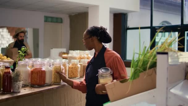 Африканский американский продавец в супермаркете заправляет полки макаронами или соусами, свежесобранными продуктами в ящиках. Владелец малого бизнеса в окрестностях нулевой экологический магазин отходов. - Кадры, видео