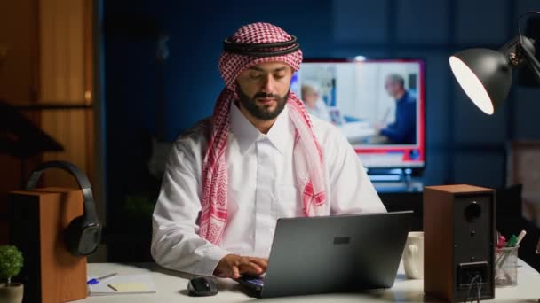 Arab ze słuchawkami bezprzewodowymi entuzjastycznie nastawiony do pracy w domu. Wesoły muzułmański biznesmen robi badania online podczas słuchania muzyki, aby czas szybciej mija - Materiał filmowy, wideo