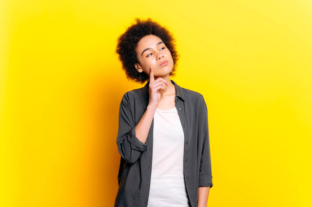 Jeune femme afro-américaine pensive aux cheveux courts et bouclés, élégamment habillée, regardant vers le côté, réfléchissant à quelque chose, planifiant, se tenant debout sur un fond jaune isolé - Photo, image