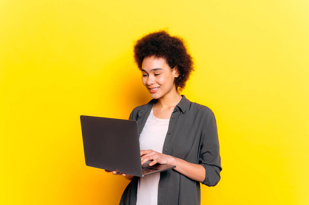 Positieve Afro-Amerikaanse of Braziliaanse jonge vrouw, vrouwelijke student, programmeur, freelancer, webdesigner, met kort krullend haar, met behulp van laptop, staat op geïsoleerde gele achtergrond, glimlachen - Foto, afbeelding