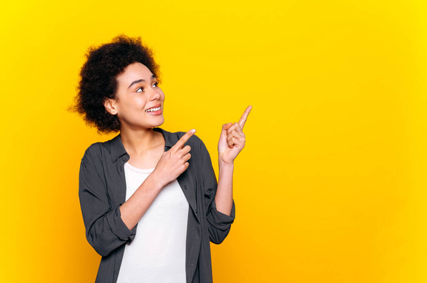 Joyful excited african american lovely curly haired woman, in casual shirt, looks and points with fingers of both hands to the side at empty space for advertise, smile, isolated yellow background - Photo, Image