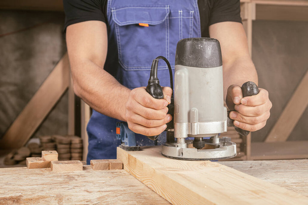 Νεαρός επαγγελματίας ξυλουργός ισούται με μια ξύλινη μηχανή άλεσης σε ένα ξύλινο τραπέζι στο εργαστήριο - Φωτογραφία, εικόνα