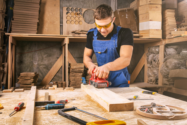Опытный плотник в рабочей одежде и владелец малого бизнеса, работающий в деревообрабатывающей мастерской, использующий наждачную бумагу для полировки дерева в мастерской   - Фото, изображение