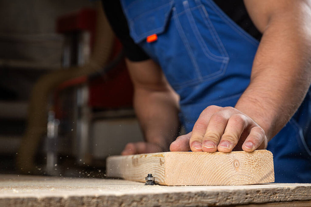 Een jonge mannelijke timmerman bouwer in het algemeen werkt gelijk aan een houten bar met een freesmachine in de werkplaats, op de achtergrond houten planken - Foto, afbeelding
