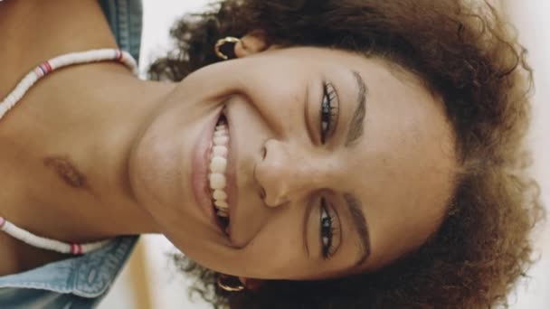Vertical médio closeup slowmo retrato de alegre jovem mulher negra sorrindo com os dentes na câmera de pé no shopping center contemporâneo em fundo borrado - Filmagem, Vídeo