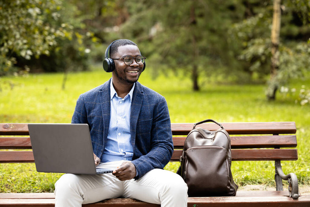 Egy férfi fejhallgatóval és laptoppal a padon ül a parkban, és az irodán kívül dolgozik.. - Fotó, kép