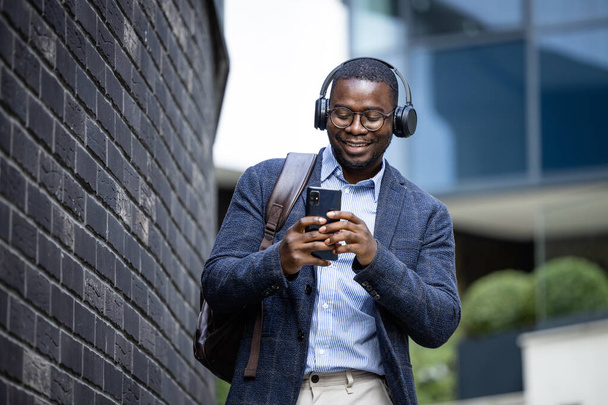 Ευτυχισμένος άντρας με κουστούμι ακούγοντας μουσική στα ακουστικά και πηγαίνοντας στη δουλειά. Στο παρασκήνιο σύγχρονα κτίρια. - Φωτογραφία, εικόνα